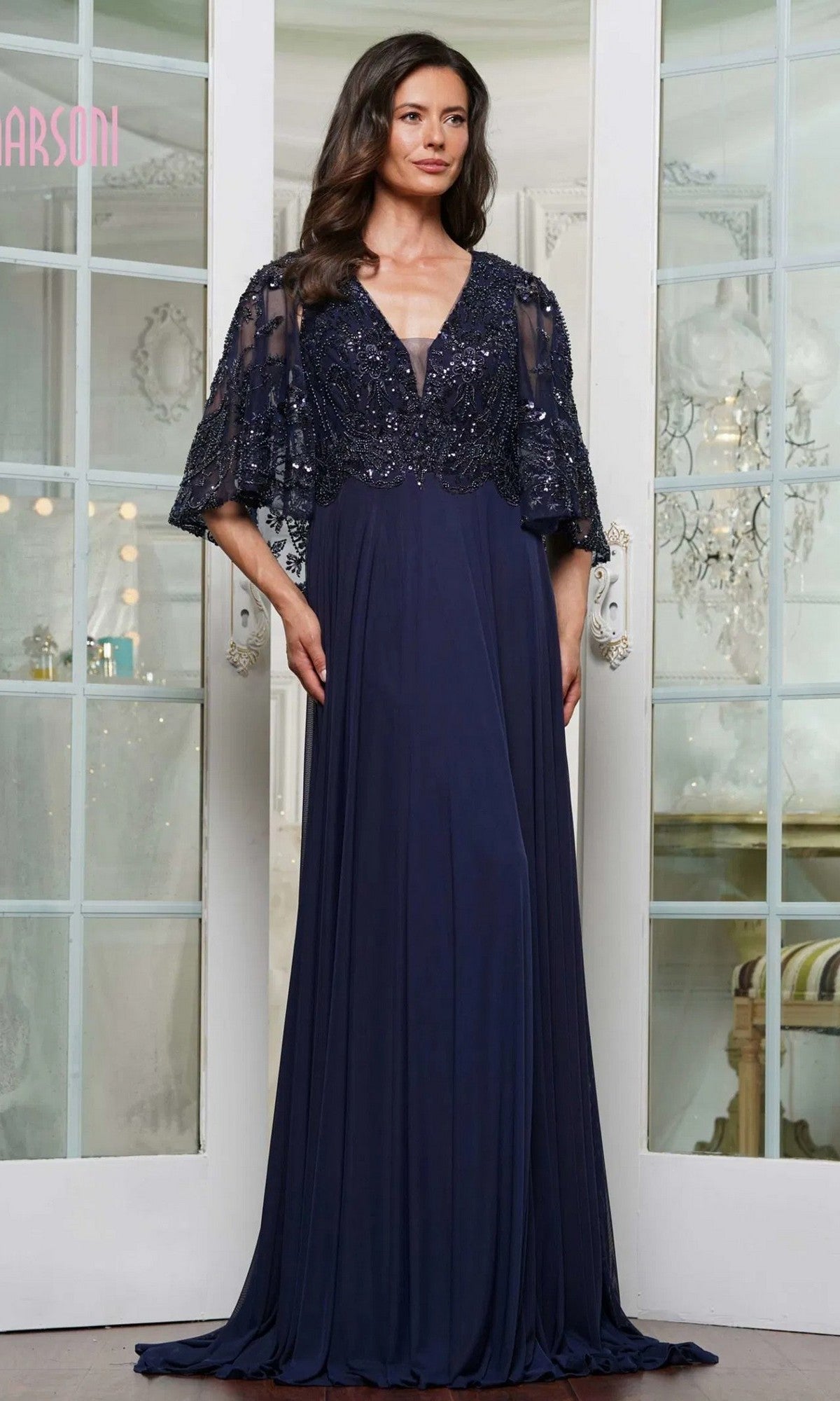A woman wearing the Marsoni MV1302 long formal dress in navy, featuring beaded details and a sheer cape overlay, stands before white French doors.